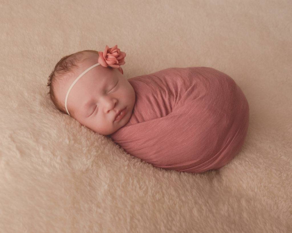 A newborn girl wrapped in a pink wrap sleeping on a fluffy blanket.
