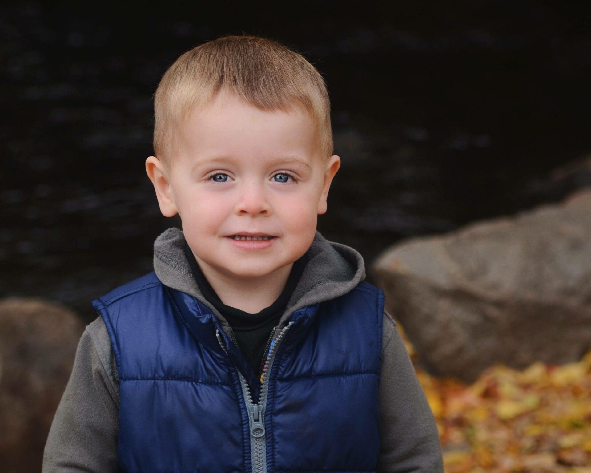 A young boy in a blue vest standing in front of rocks.