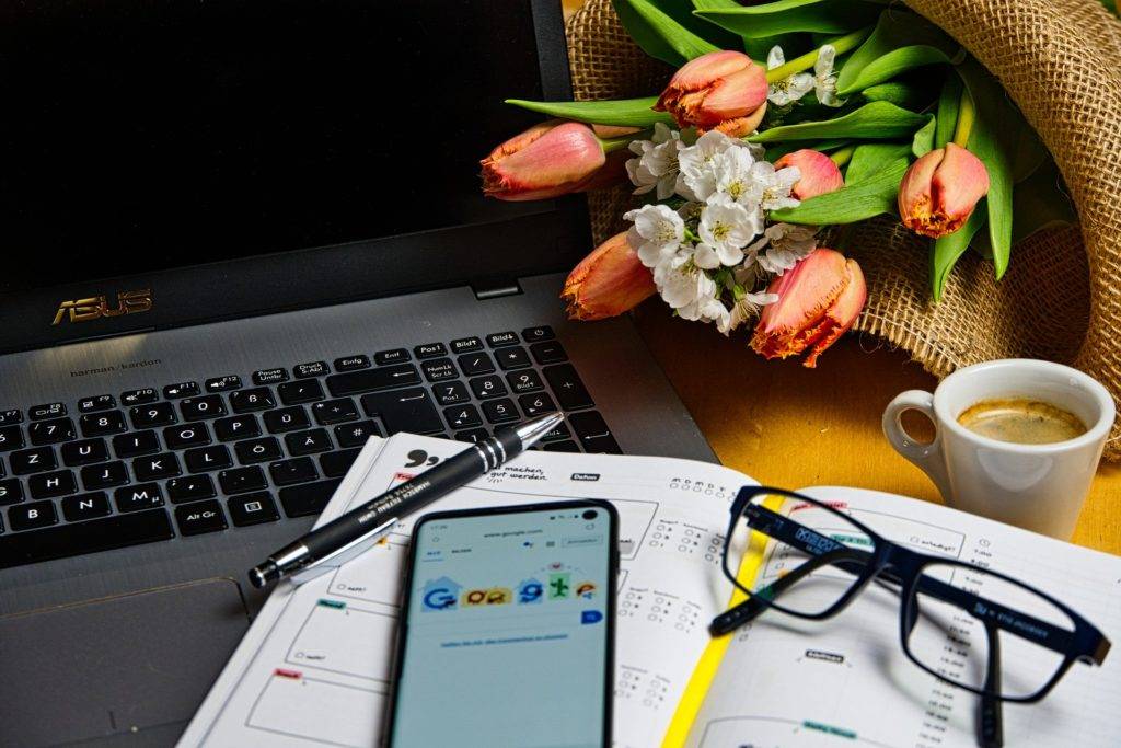 A laptop on a table next to a phone and a vase of tulips.