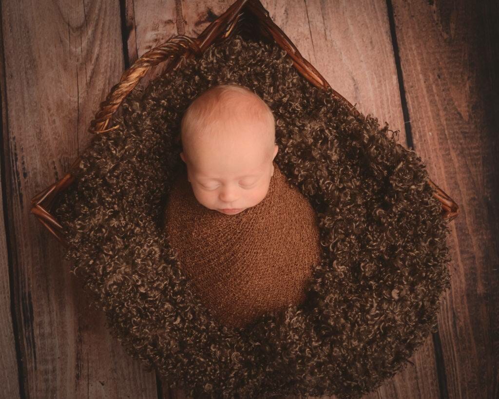 A baby sleeping in a brown basket on a wooden floor.
