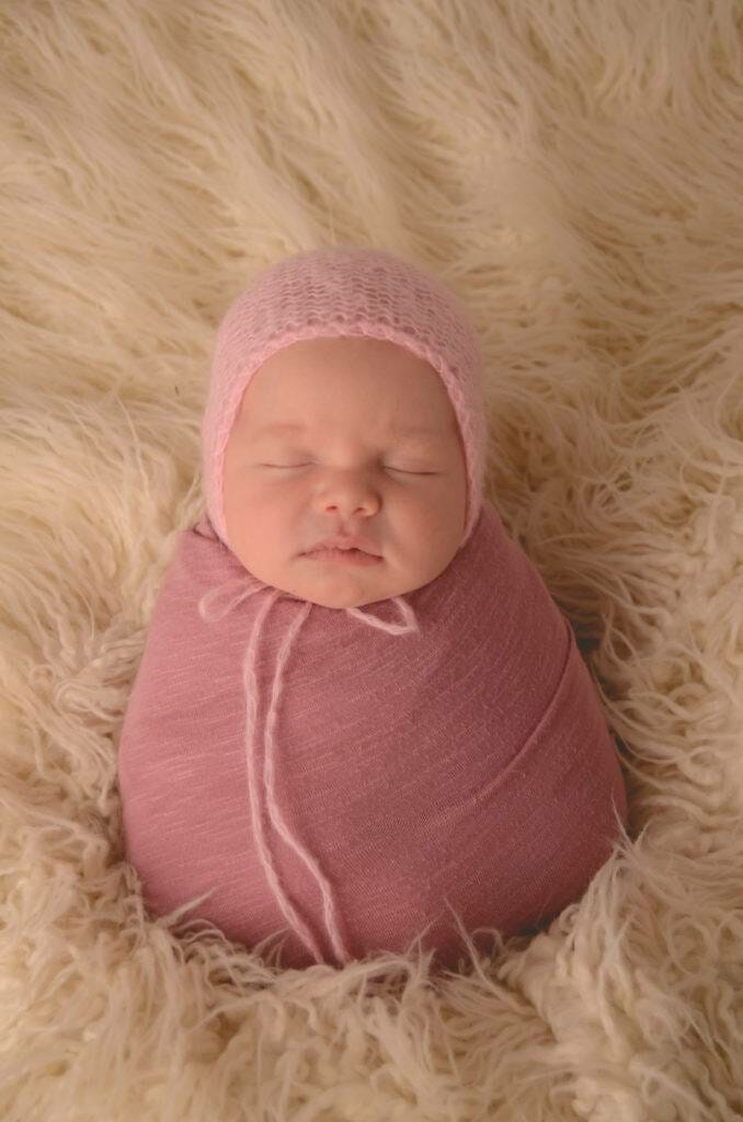 A baby in a pink hat laying on a fluffy blanket.
