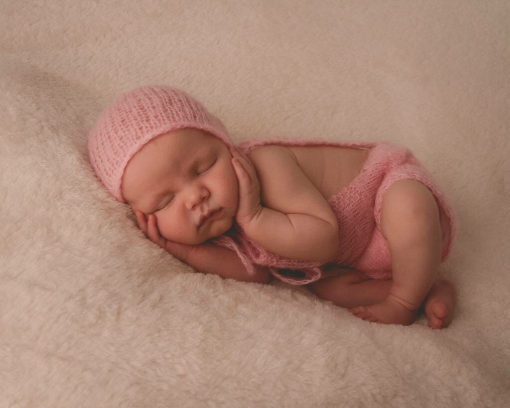 A baby girl in a pink knitted outfit laying on a white blanket.