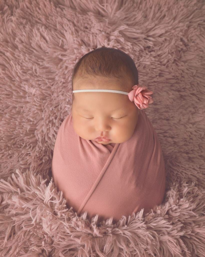 A baby girl wrapped in a pink blanket on a fluffy rug.