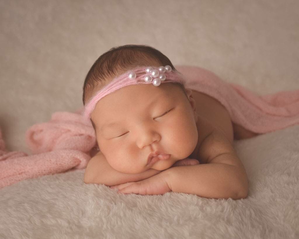 A baby girl wearing a pink headband is laying on a blanket.