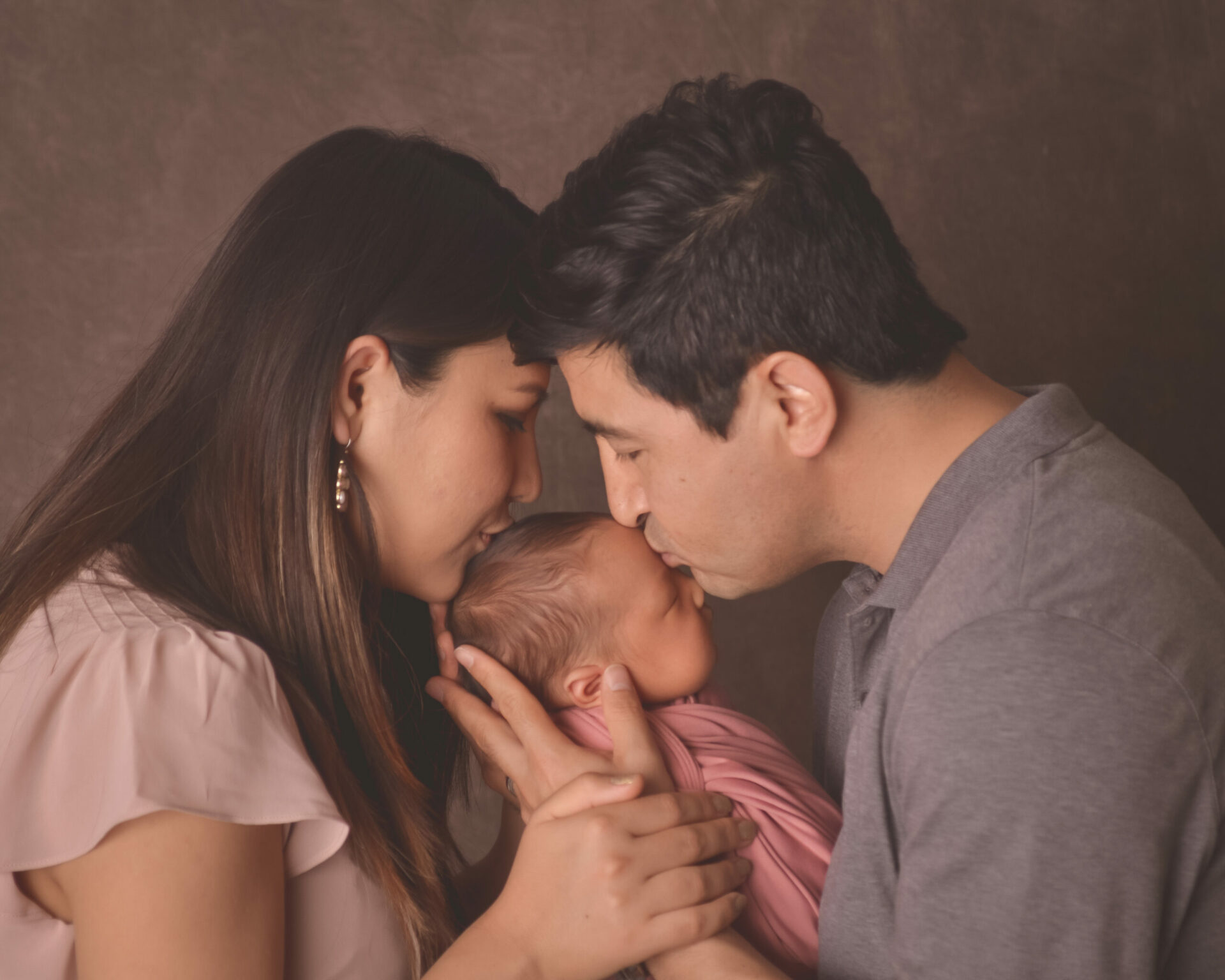 A man and woman kissing their baby in front of a gray background.