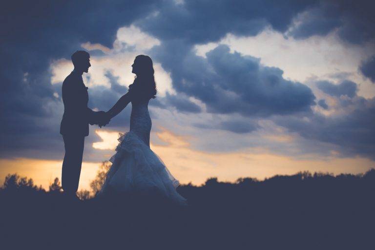 A bride and groom are silhouetted against a cloudy sky.