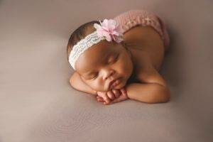 A baby girl is laying down with a pink flower headband on her head.