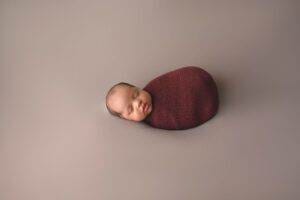 A newborn wrapped in a burgundy blanket on a gray background.