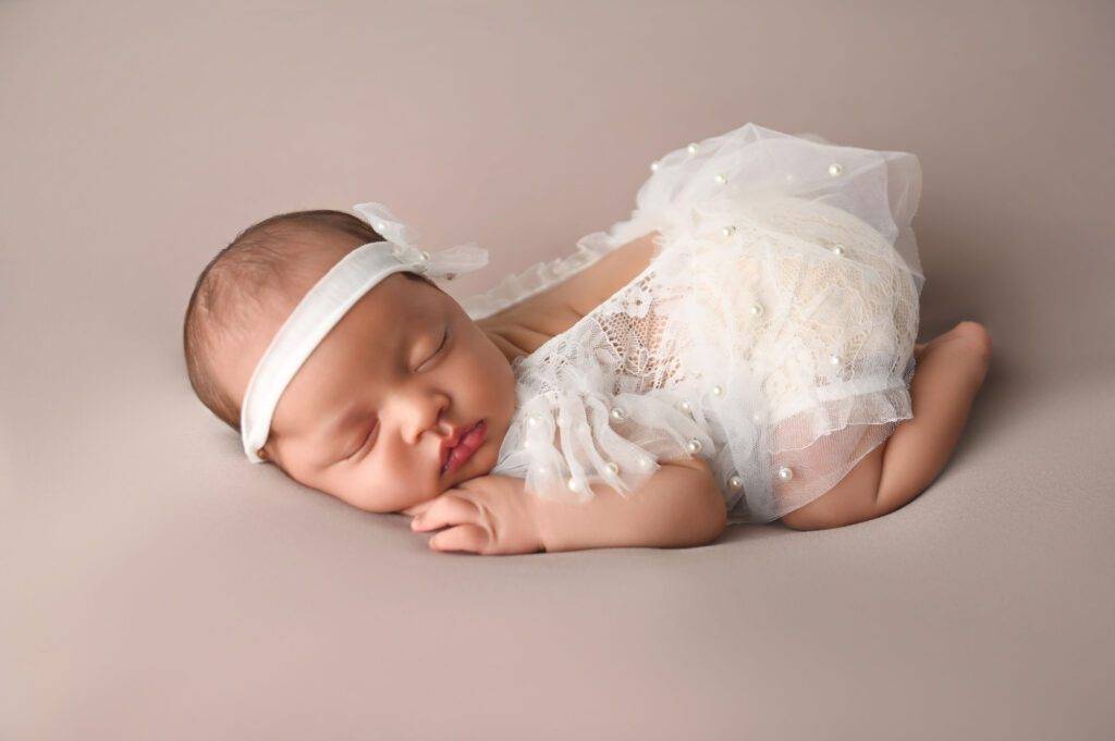 A baby girl in a white outfit is laying on a grey background.