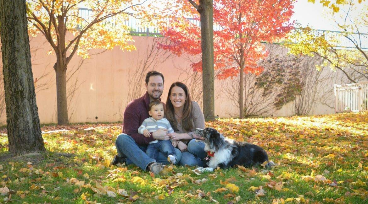 A family sits on the ground with their dog in the fall leaves.