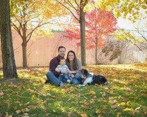 A family sits on the ground with their dog in the fall leaves.