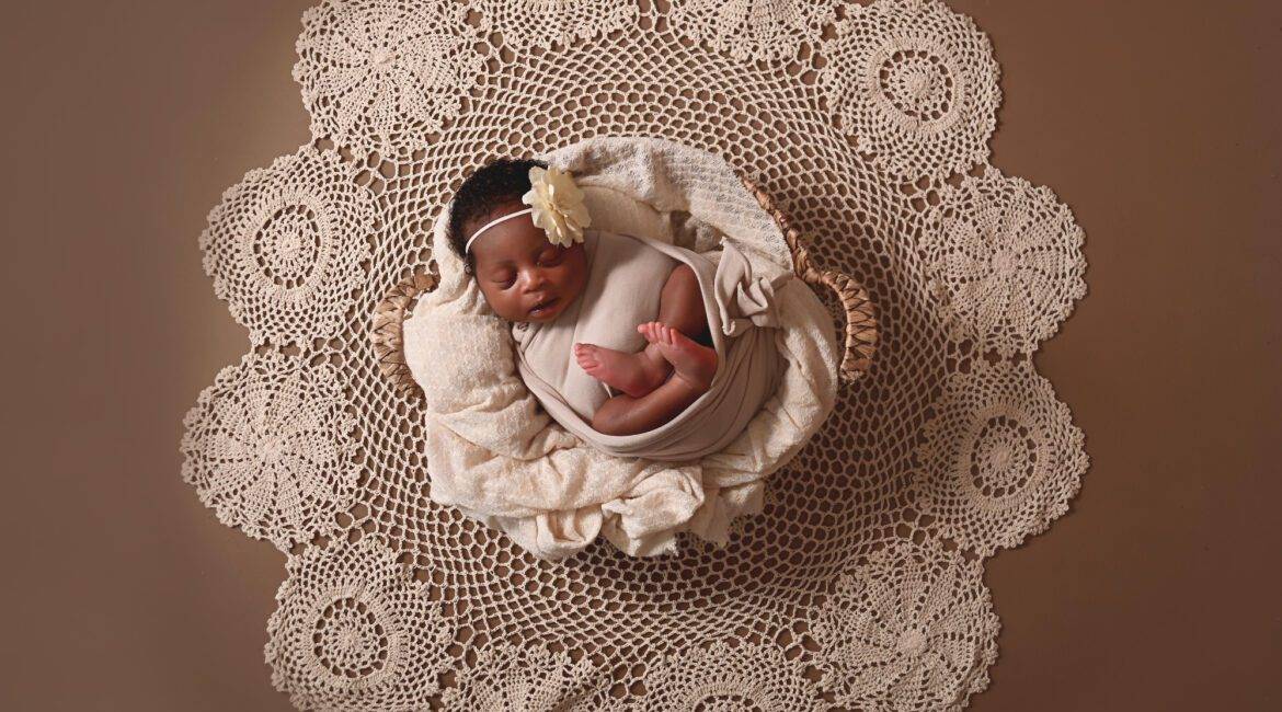 A newborn baby laying on a doily on a brown background.