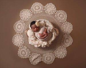 A newborn baby laying on a doily on a brown background.