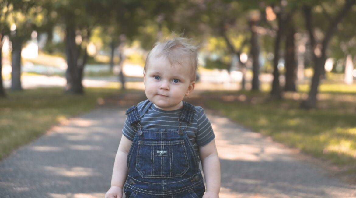 A baby boy in overalls standing on a path.
