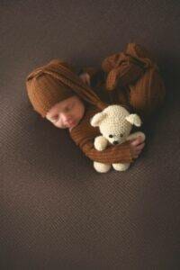 A newborn baby laying on a brown blanket with a teddy bear.