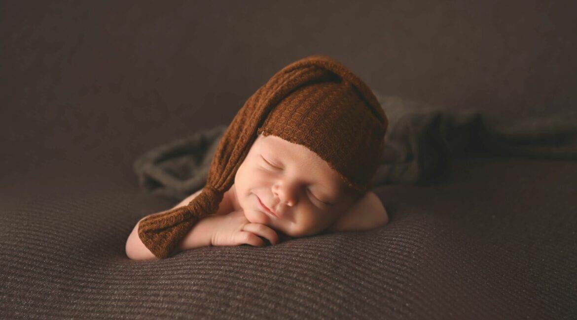 A baby wearing a brown knitted hat is laying on a blanket.