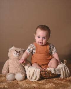 a baby sitting on a basket holding a teddy bear