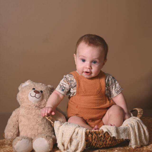 a baby sitting on a basket holding a teddy bear