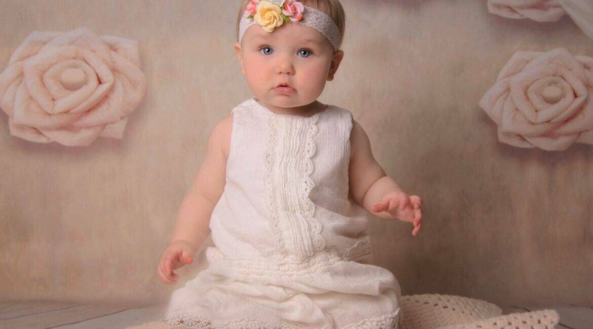 A baby in a white dress sitting on a blanket.