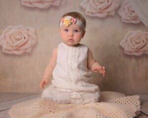 A baby in a white dress sitting on a blanket.