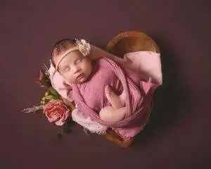 A newborn girl in a pink wrap laying in a heart shaped bowl.