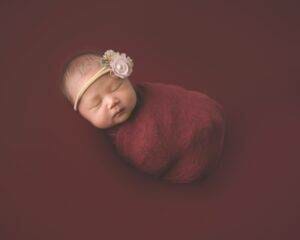 A newborn girl wrapped in a red blanket with a flower on her head.
