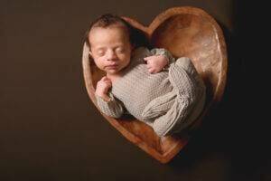 A baby boy is laying in a wooden heart shaped bowl.