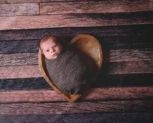 A baby boy in a heart shaped bowl on a wooden floor.