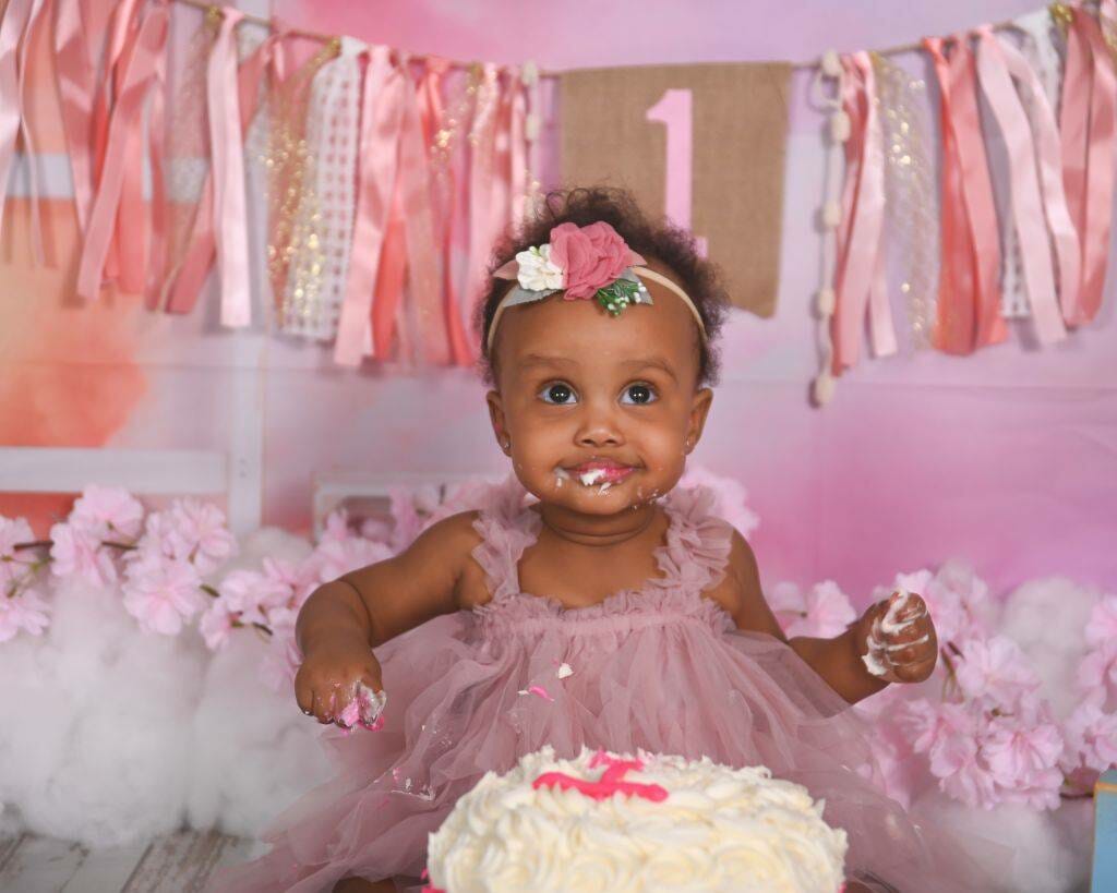 A baby girl is sitting in front of a pink cake.