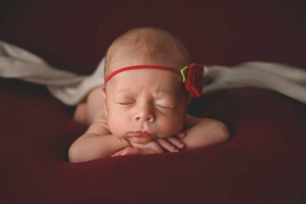A baby girl wearing a red flower headband is laying on a red blanket.