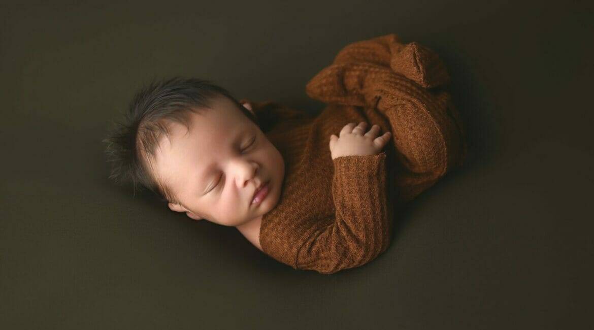 A baby boy in a brown sweater laying on a dark background.