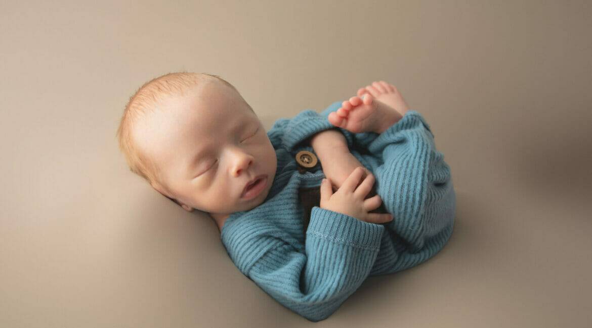 A baby boy in a blue sweater is laying on a beige background.