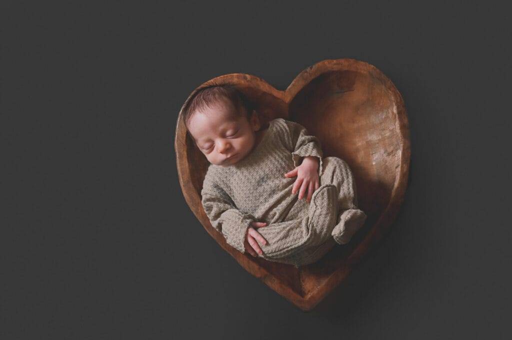 newborn in a heartbowl, wearing grey pajamas on a grey background.