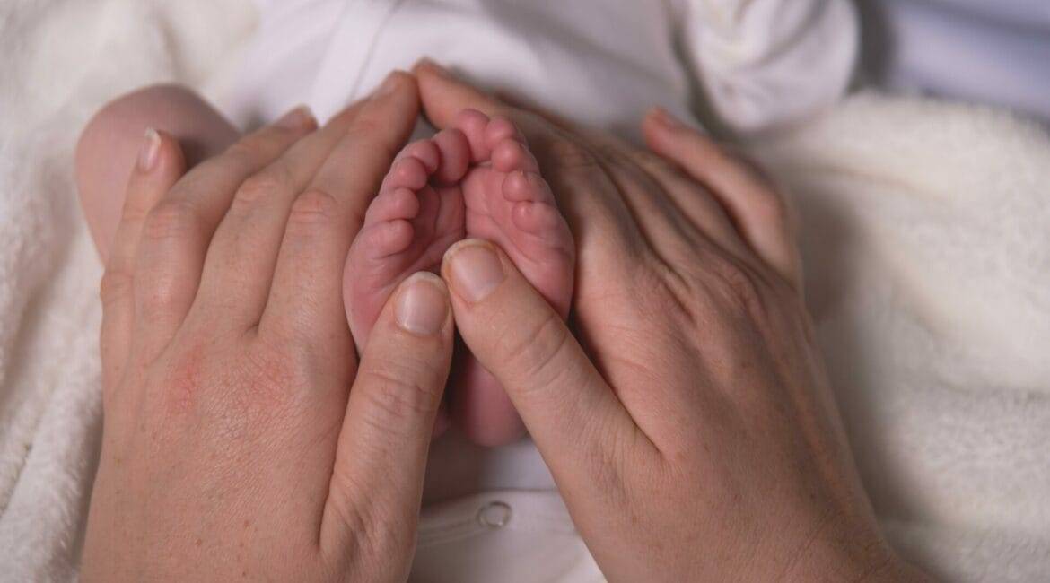 "The image captures a tender moment where an adult's hands gently cradle a newborn baby's tiny feet. The baby's feet are small and delicate with little toes, highlighted against the adult's larger, caring hands. The baby appears to be lying on a soft, white blanket, with a glimpse of a white onesie visible. The scene conveys a sense of warmth, care, and the intimate bond between parent and child.