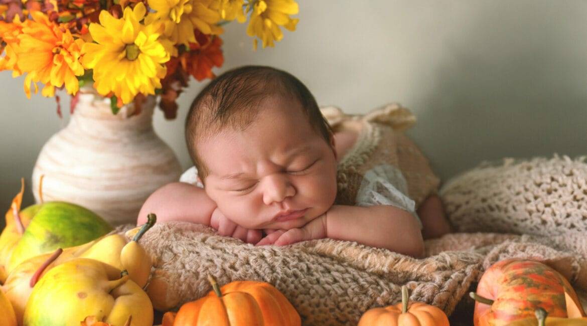 Newborn autumn photography Baby wrapped in burgundy cloth Fall-themed newborn portrait Infant pumpkin photo session Cozy autumn baby picture Newborn photography with fall props Baby in seasonal setup Autumnal newborn photoshoot Warm-toned newborn photography Festive baby portrait