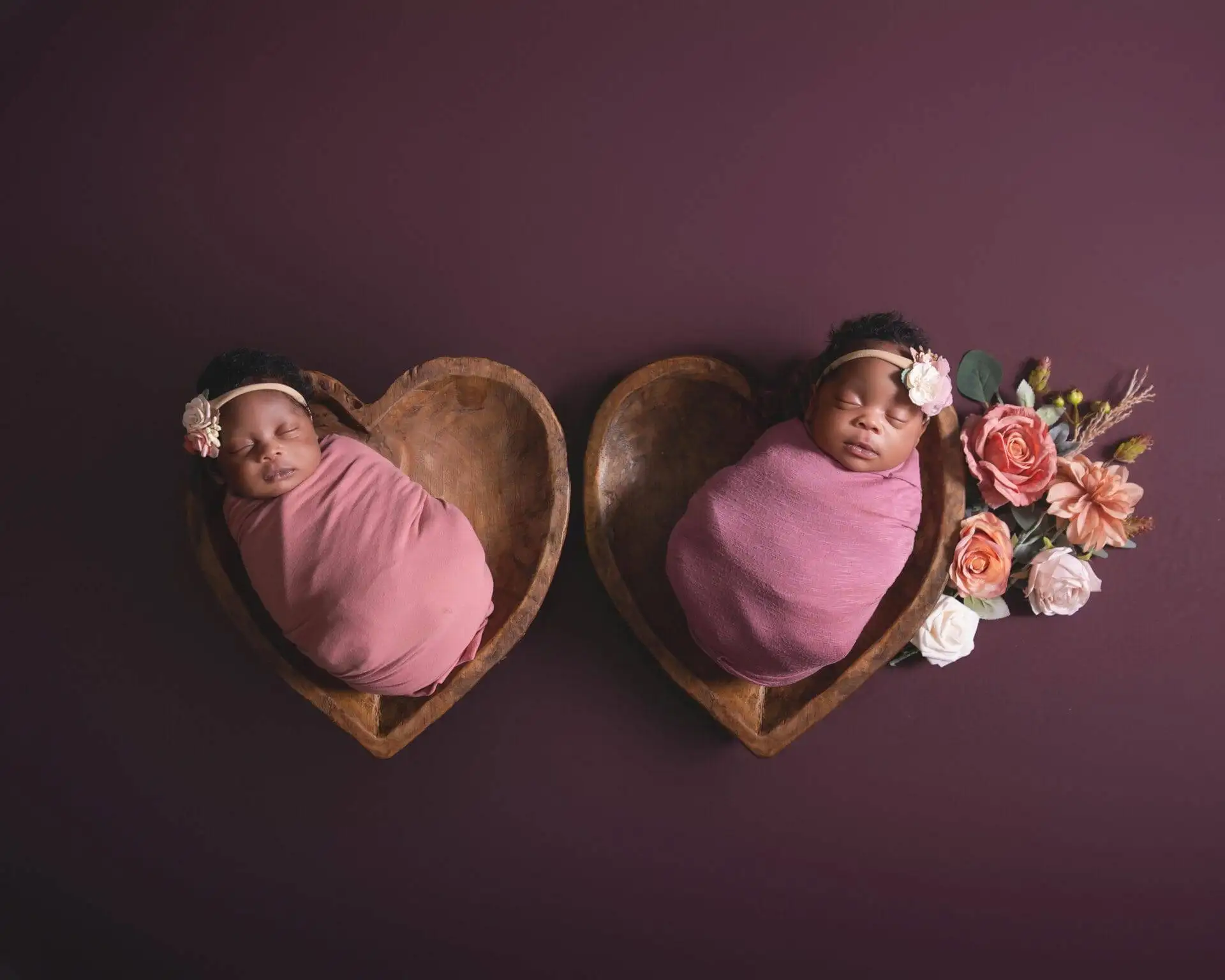 Two adorable newborn babies peacefully nestled in heart-shaped wooden bowls, beautifully adorned with delicate floral accents.