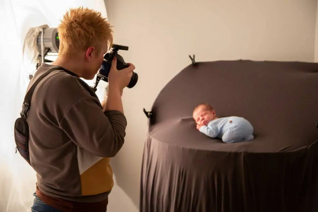A photographer with a camera captures a photo of a newborn baby swaddled in blue, lying on a brown circular backdrop.