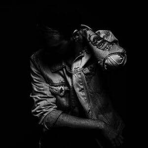 A black and white photo of a person in a denim jacket, holding their hand to their forehead, against a dark background.