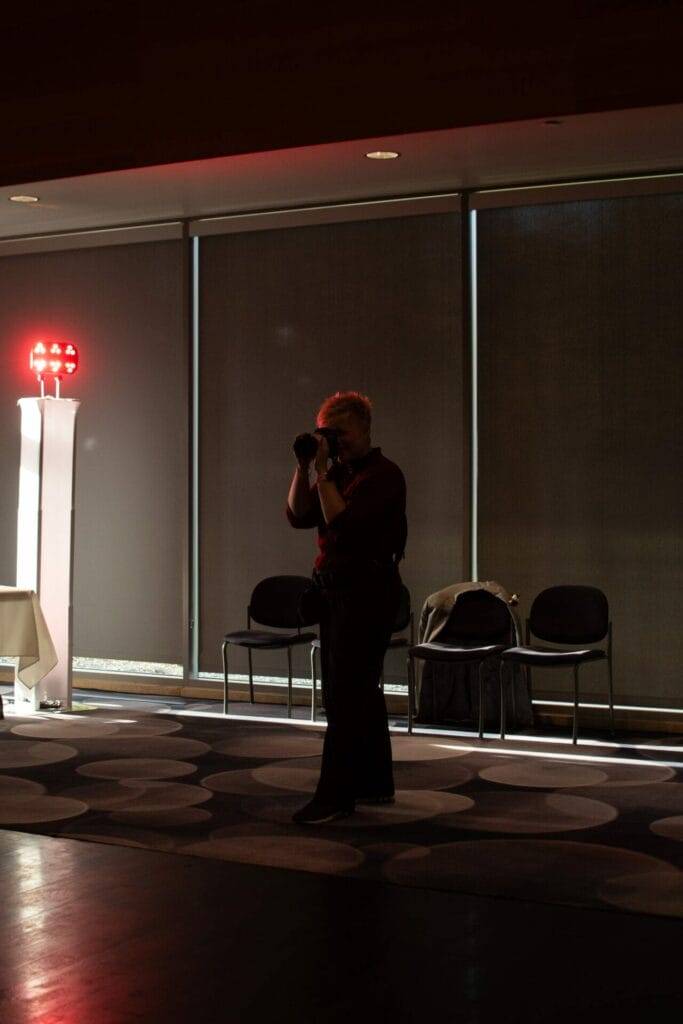 A person standing indoors taking a photo with a dslr camera, backlit by natural light from large windows, with chairs and a decorative lamp in the background.