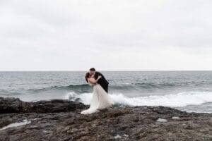 A bride and groom kissing on a rocky shore with waves crashing behind them under an overcast sky.