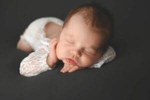 Sleeping baby in white lace outfit lying on a dark surface with head resting on hands.