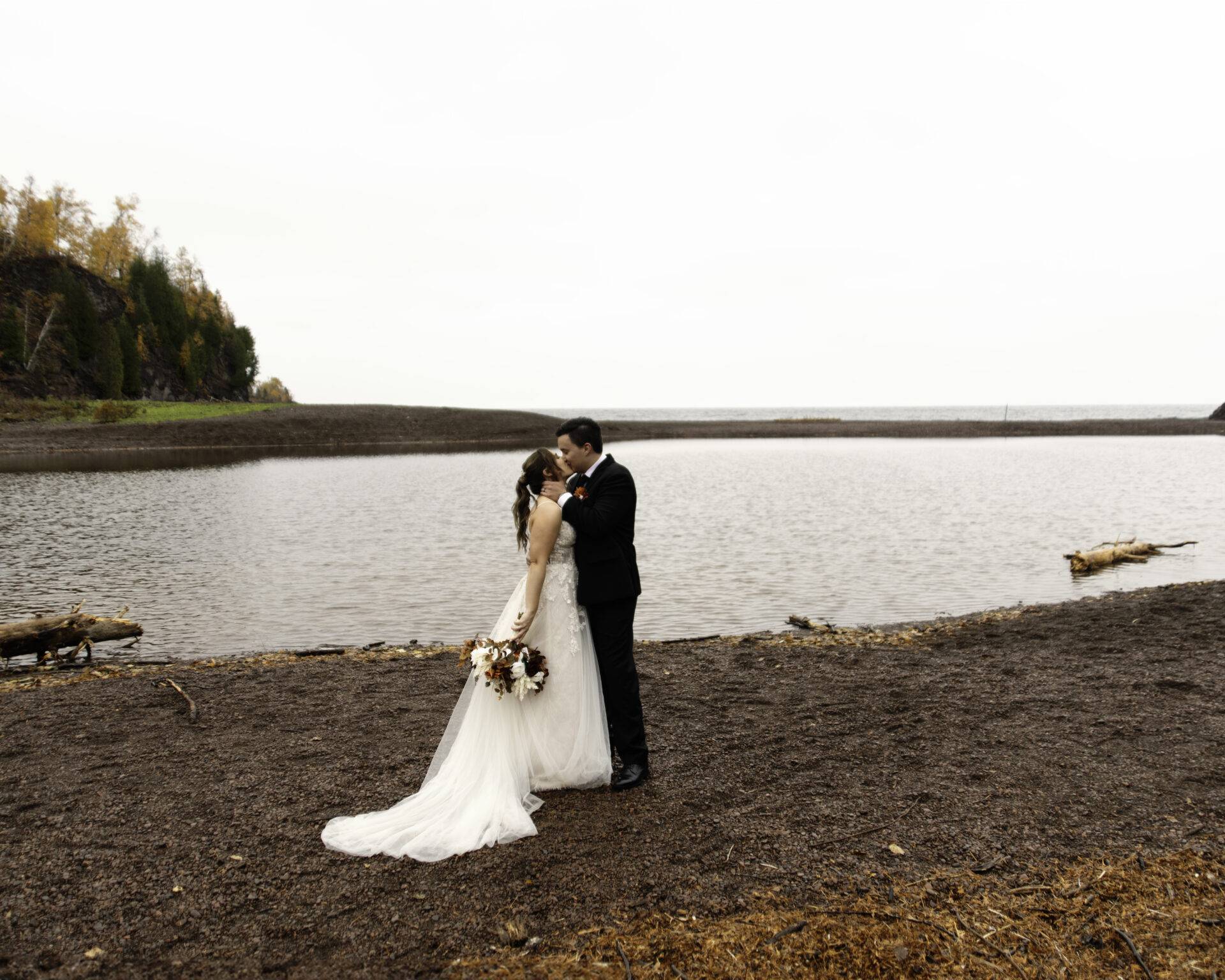 A bride and groom kissing on a sandy lakeshore, the bride holding a bouquet, with a forested hill in the background.
