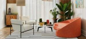 A modern living room with a large window, armchairs, a round coffee table, a orange sofa, plants, a striped rug, abstract art, and a wooden cabinet with a sculpture and table lamp.