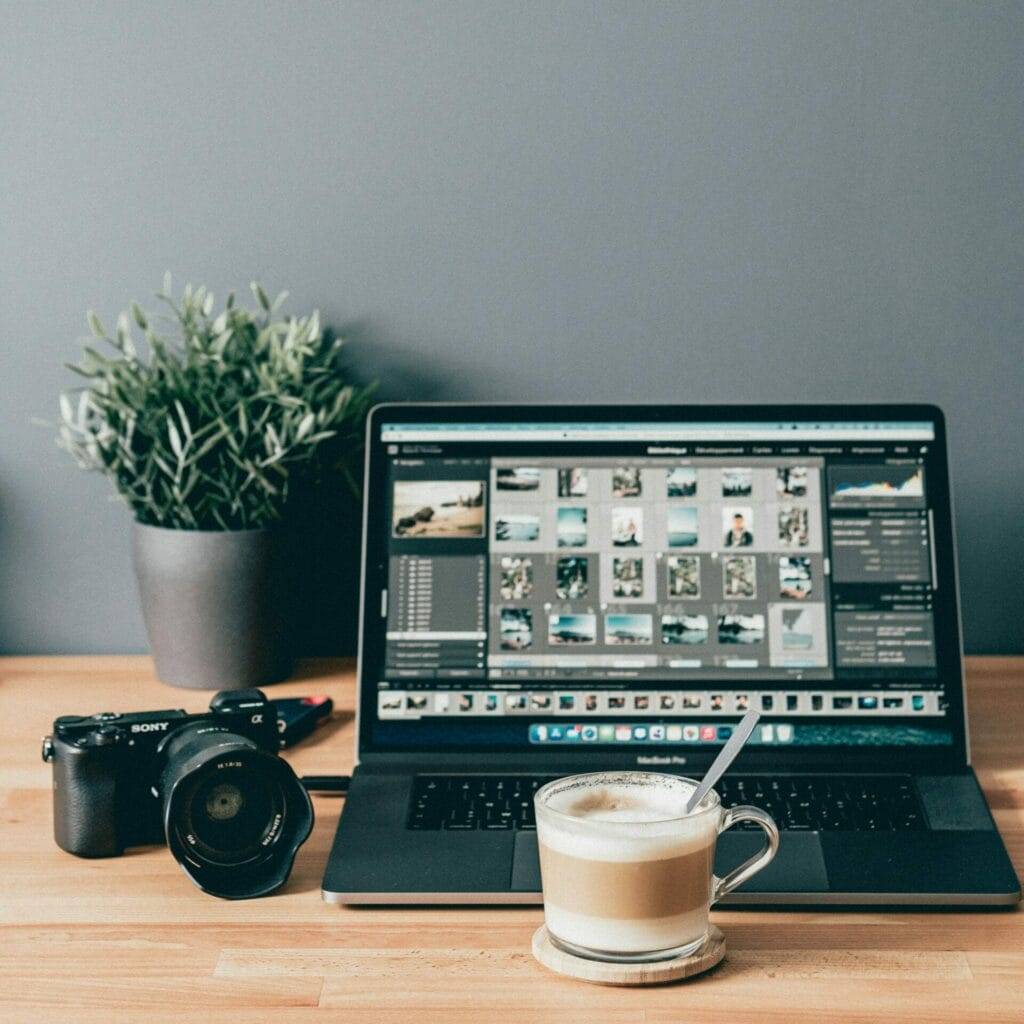 A laptop displaying photo editing software, a camera, a plant, and a cup of coffee on a wooden desk.