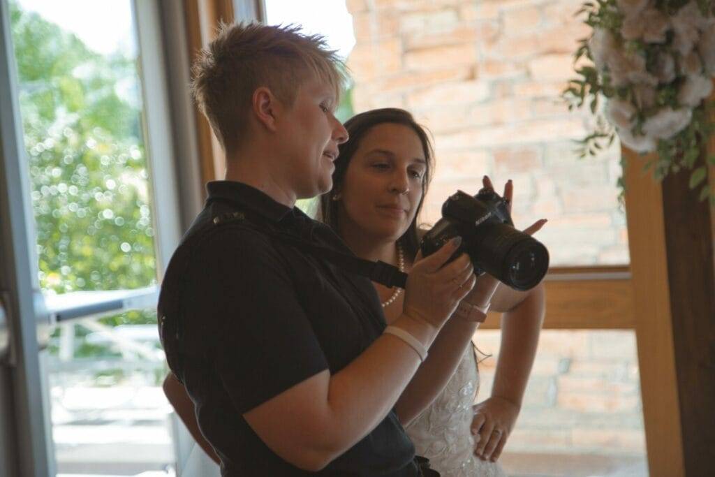 Giliane Mansfeldt and bride look at a camera's screen, reviewing photos. One person holds the camera while the other, standing close, observes attentively. They are indoors, with natural light coming through a window.