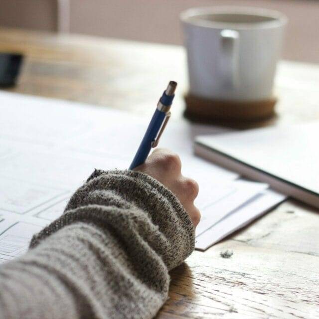 A person is writing on papers on a wooden table with a blue pen. A white ceramic mug and a white notebook with a pen lie adjacent to the papers.