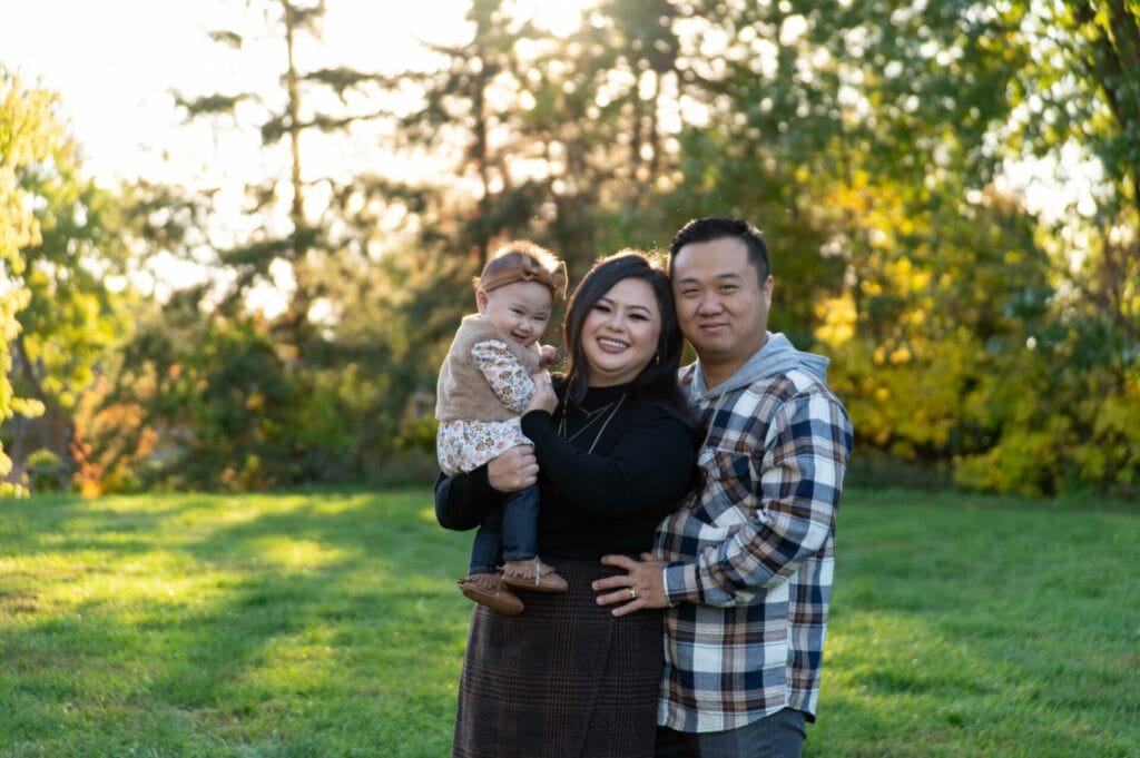 A family of three poses outdoors, with the woman holding a child, and the man standing to her right. Trees and grass fill the background.