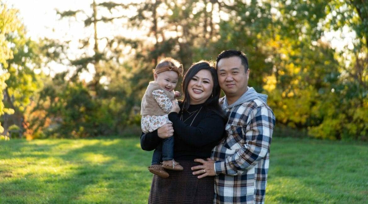 A family of three poses outdoors, with the woman holding a child, and the man standing to her right. Trees and grass fill the background.