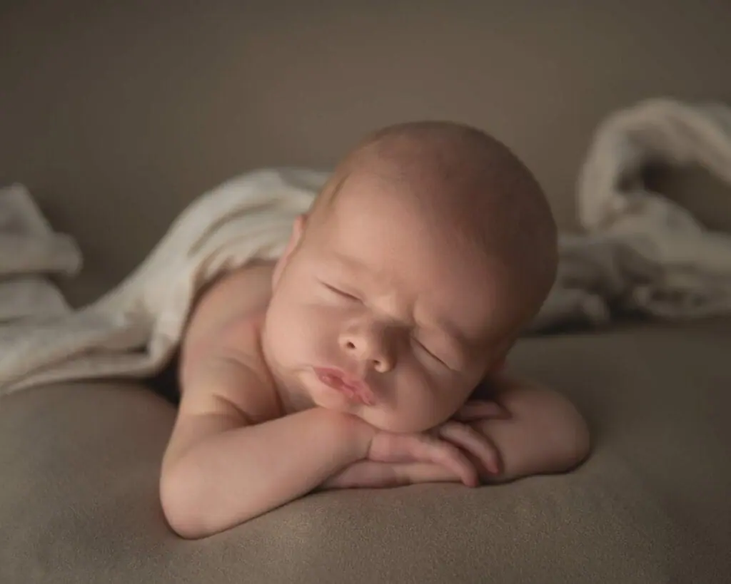 A newborn baby sleeps on its stomach with a blanket partially covering its body. The baby has its head resting on its crossed arms.