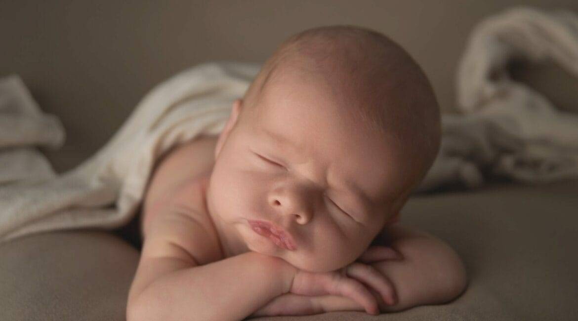 A newborn baby sleeps on its stomach with a blanket partially covering its body. The baby has its head resting on its crossed arms.